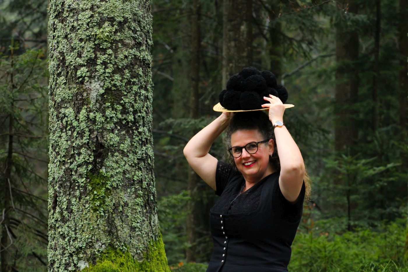 Eine Frau im Wald, sie hebt ihren Hut und schaut in die Kamera